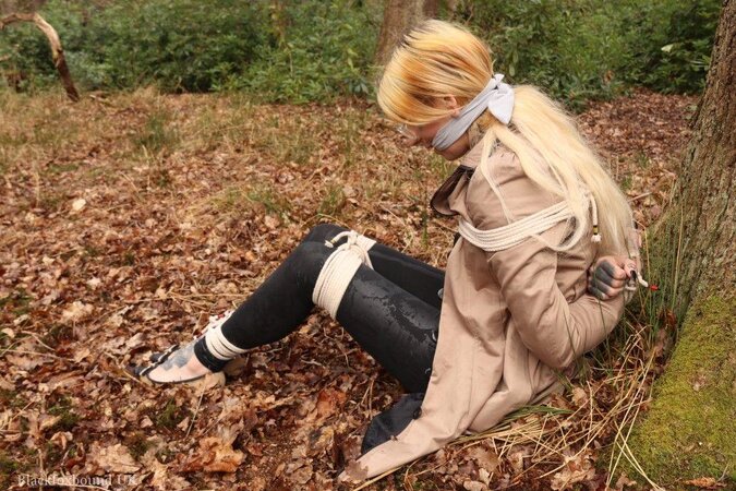Girl with glasses can't stand up because she is hogtied by the tree